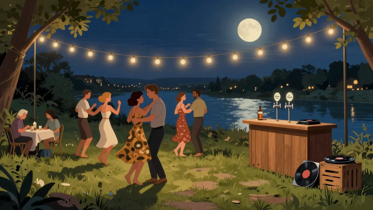 People dancing barefoot outdoors under string lights beside a river, with a simple bar and vinyl DJ setup.